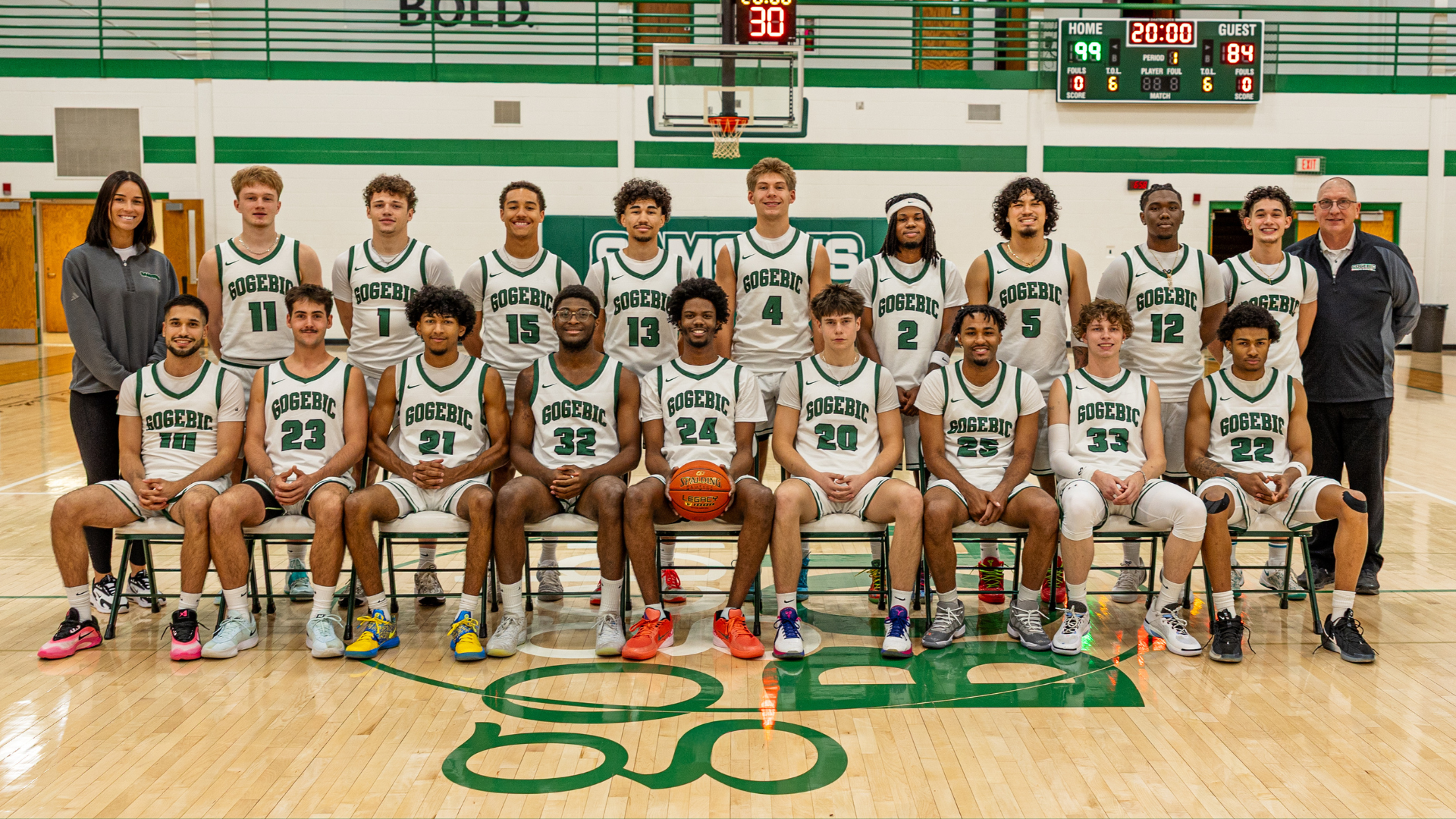 Team picture in the gym of men's basketball players and coaches, one row sitting and the back row standing.