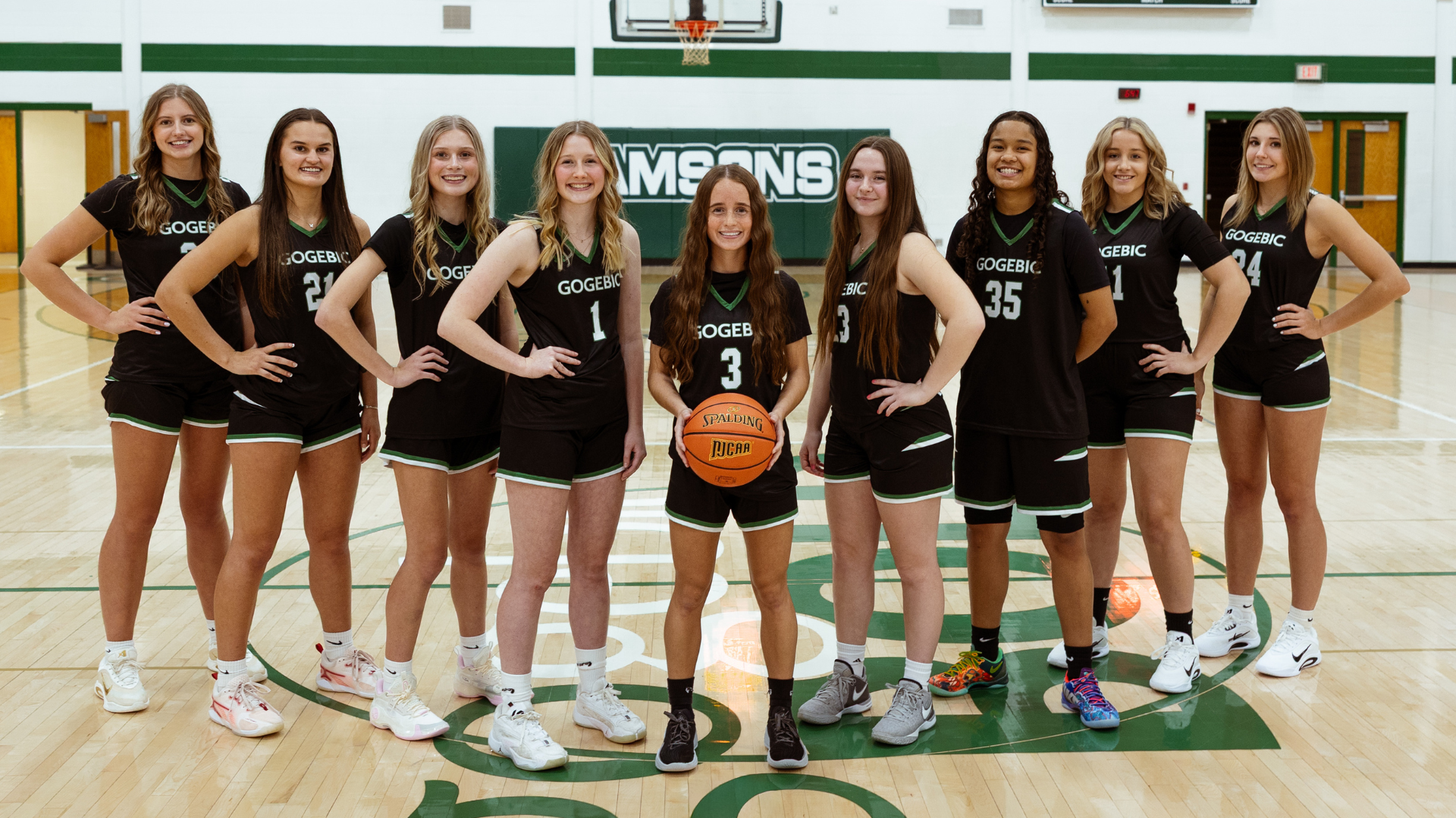 Women's basketball team lined up, holding a basketball, standing in the gym.